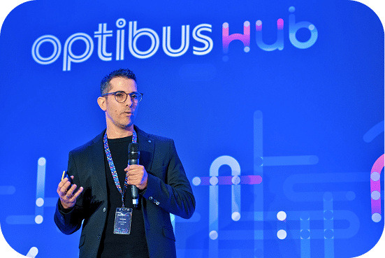 A speaker presents at the Optibus Hub conference against a vibrant blue backdrop, gesturing while holding a microphone and wearing a credential badge.