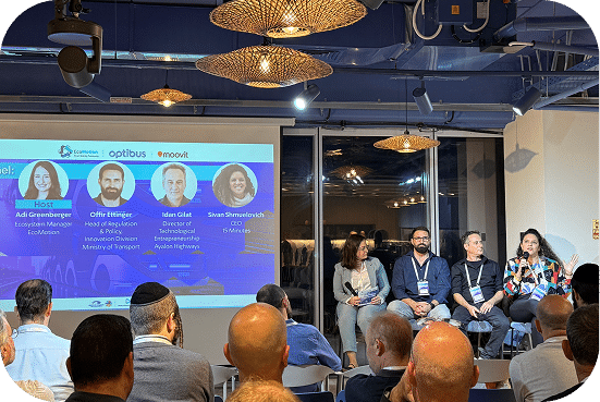 Professional conference panel discussion with five speakers on stage addressing an engaged audience, featuring a digital display showing panelist names and organizational affiliations in a