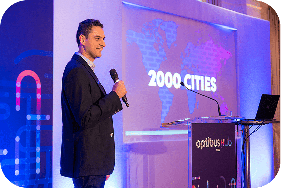 Professional speaker in business attire presents at a tech conference, discussing global expansion to 2000 cities with a world map displayed behind him.