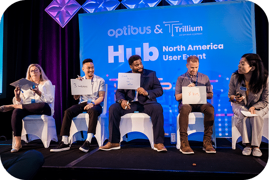 Five panelists sit on stage at the Optibus and Trillium North America Hub User Event, each holding laptops while discussing industry topics in front of a blue branded backdrop.
