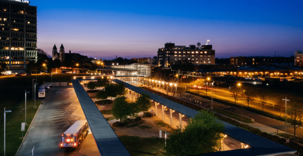 Urban riverside park at twilight features illuminated walkways, historic church spires, and city buildings glowing against a deepening purple sky.