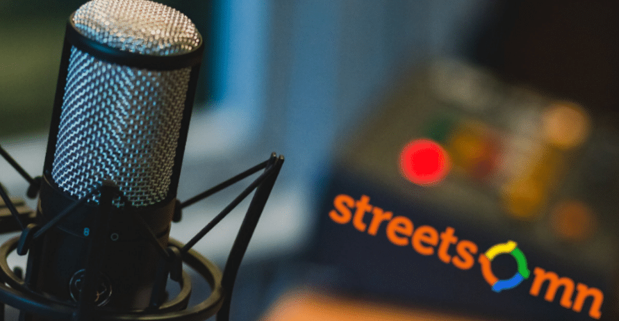 Professional studio microphone with blue mesh grille mounted on stand, with blurred Streetcomm branding visible in the background, suggesting podcast or broadcasting equipment setup.