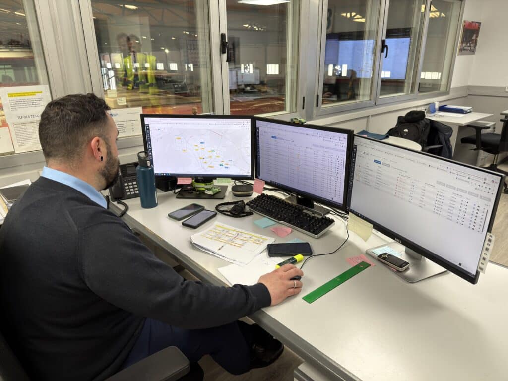 Professional working at a desk with three monitors displaying network maps and data spreadsheets in a modern office with floor-to-ceiling windows overlooking a cityscape.