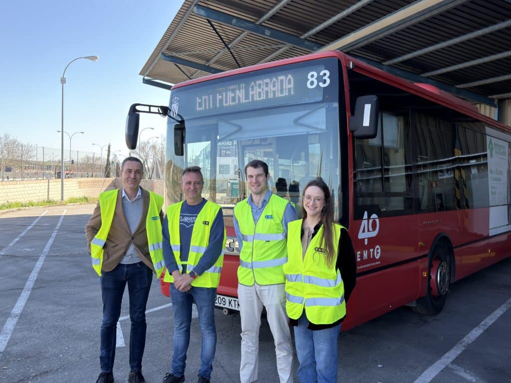 Four professionals wearing safety vests stand in front of a red bus numbered 83 at a transit station on a clear day.