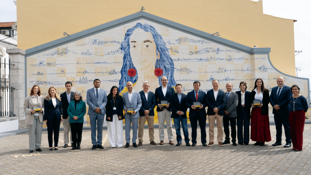 Group of business professionals pose together in front of a large mural featuring a woman's face with blue hair and cityscape backdrop. Several hold HERO awards.