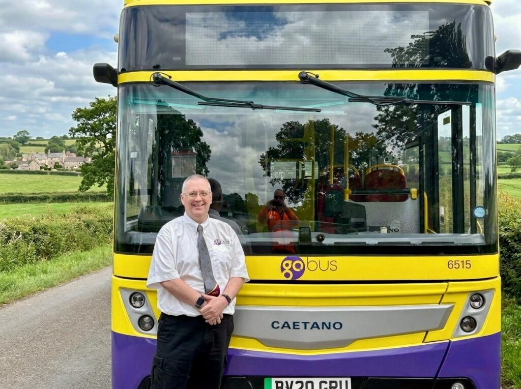 Bus driver poses in front of a yellow and purple Go Bus coach numbered 6515 in rural English countryside with green fields and village buildings visible behind.