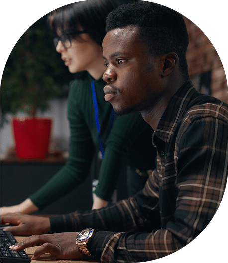 Two people work together at a computer, with the man in a plaid shirt focused on the screen while his colleague assists nearby.