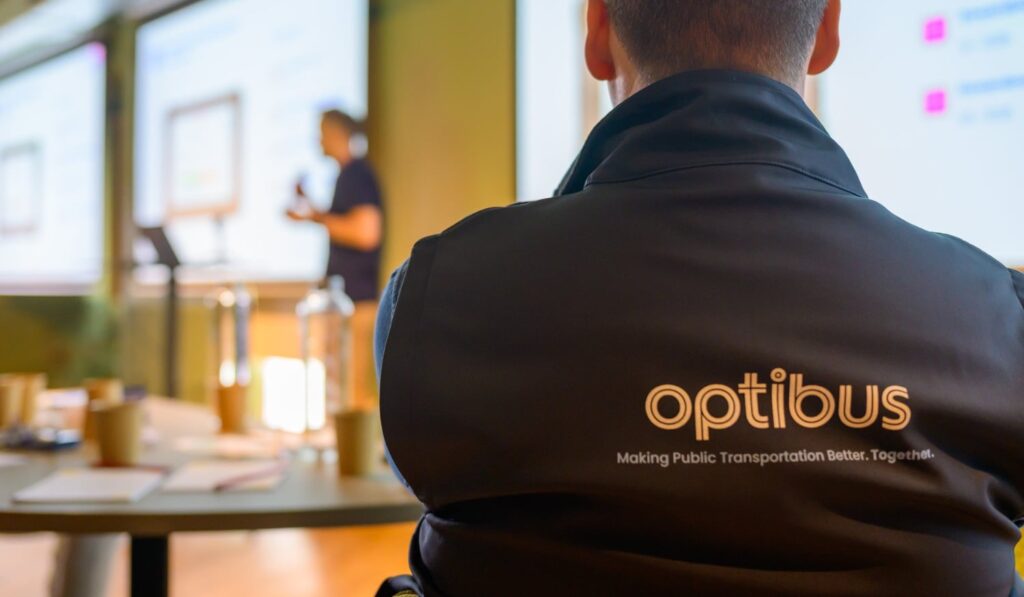 Person wearing Optibus branded jacket at a public transportation industry event, with presentation screens visible in the background during a business meeting.
