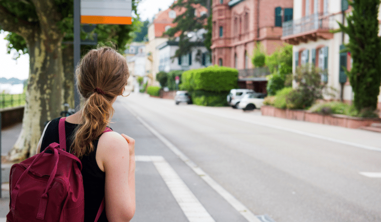 Young traveler with burgundy backpack stands on a tree-lined European street, gazing toward charming red-brick buildings and parked cars in a picturesque town setting.
