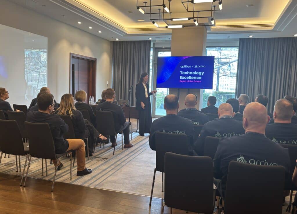Business conference presentation on technology excellence featuring a female speaker addressing seated attendees wearing Arriva branded apparel in a modern hotel ballroom.