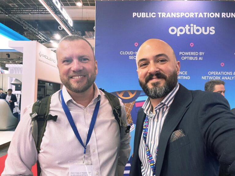 Two professionals wearing conference badges pose at an optibus public transportation event, smiling before a blue branded backdrop highlighting cloud-native solutions and AI-powered operations