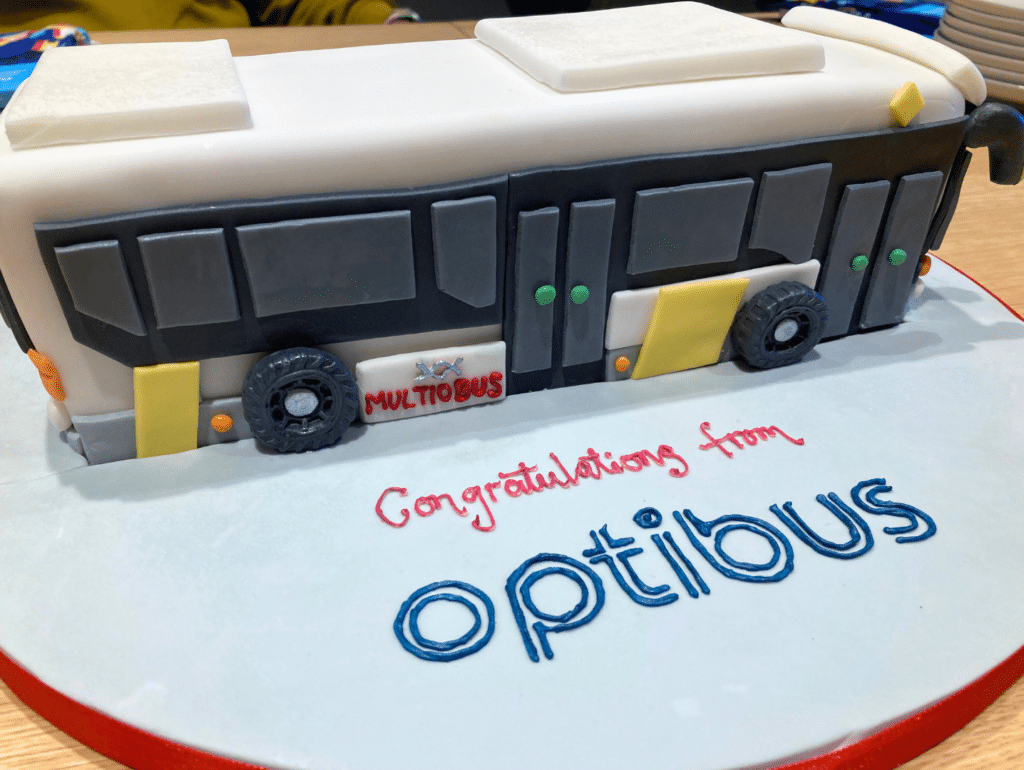 A decorative bus-shaped cake congratulates Optillous with white fondant, yellow and black details, and dark wheels. Red piping reads "Congratulations from" above the blue Optillous logo on the