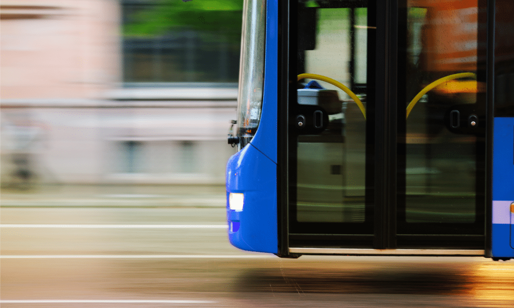 A blue public bus in motion captures motion blur on the street, showing open doors with yellow handles and dark windows reflecting the urban surroundings during transit.