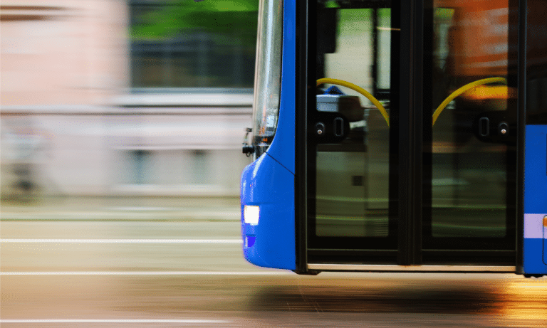 A blue public bus in motion captures motion blur on the street, showing open doors with yellow handles and dark windows reflecting the urban surroundings during transit.