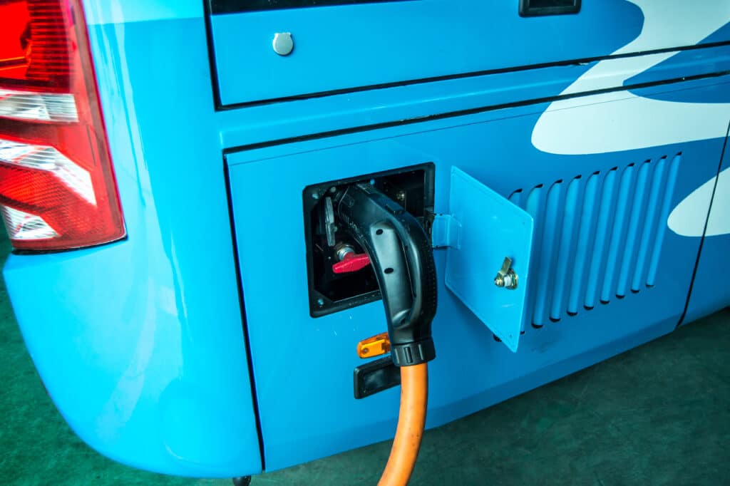 Blue electric vehicle charging with orange cable plugged into port. Modern EV with red taillights and ventilation grilles parked on green surface.