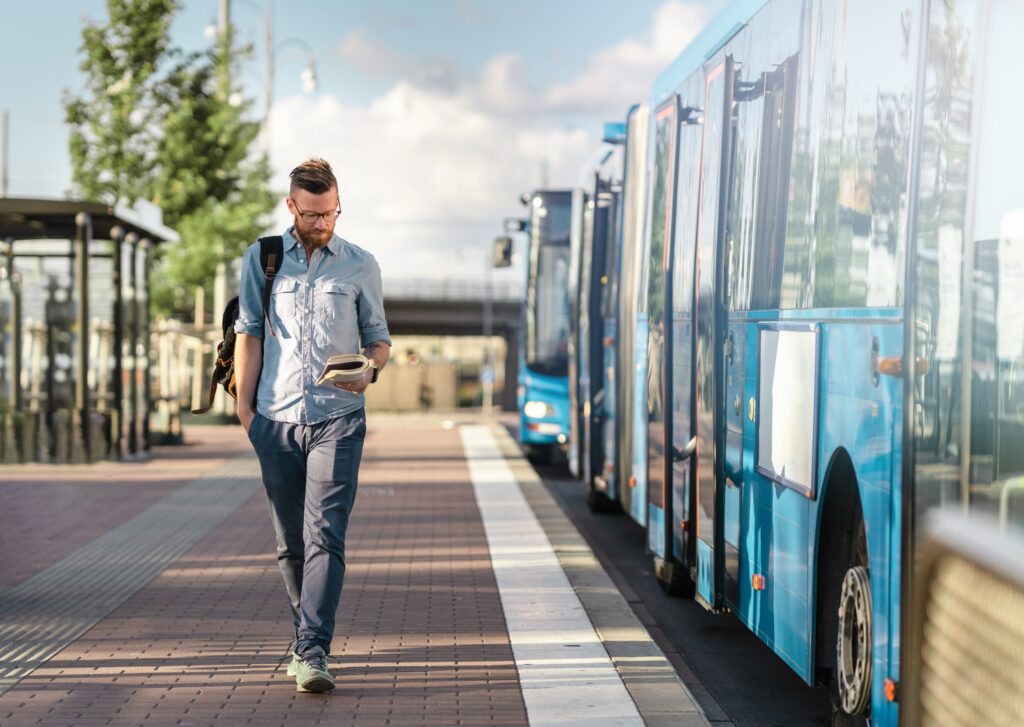 Man with glasses and backpack reading while walking past blue buses at a transit station on a sunny day.