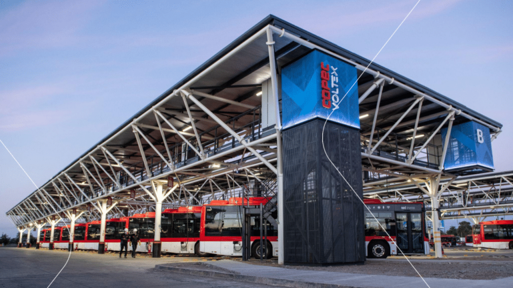 Modern bus transit station with red and white articulated buses parked under a spacious steel canopy structure, featuring blue digital signage and a black control tower.