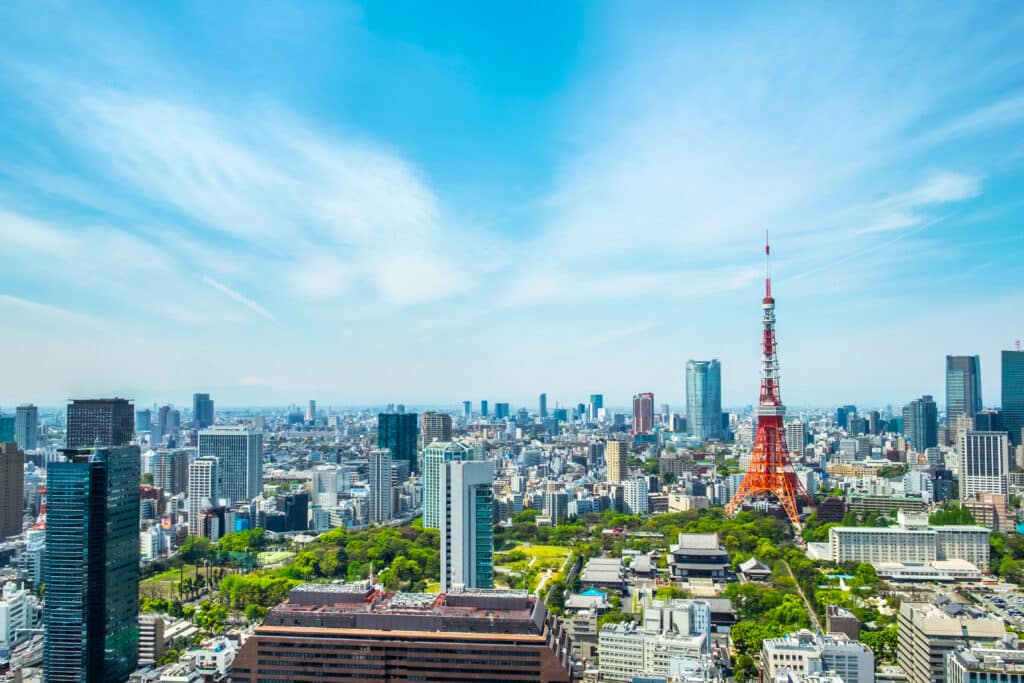 Tokyo skyline featuring the iconic red and white Tokyo Tower surrounded by modern skyscrapers, green spaces, and clear blue sky on a sunny day.