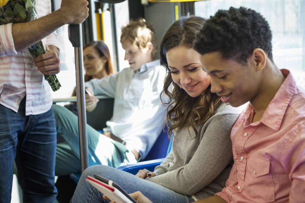 Young people on a bus reading together, smiling while holding books and documents. A man holds flowers nearby as other passengers sit in the background.