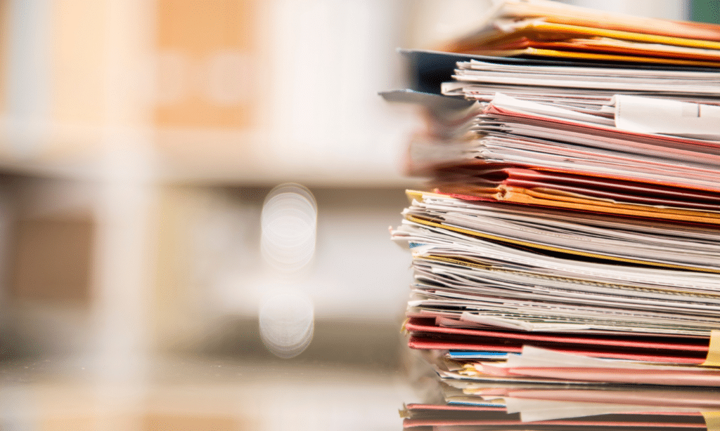 A tall stack of magazines and folders with colorful covers sits on a desk, with a blurred office background and bright window creating depth in the composition.