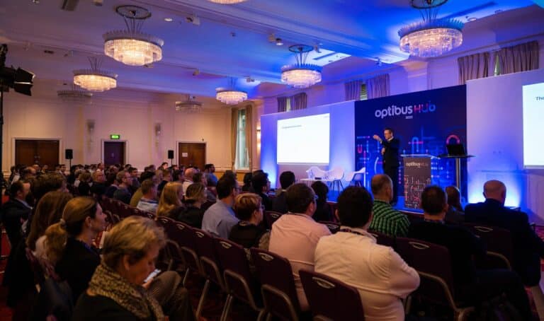Professional conference presentation at OptiBus Hub 2023 with speakers addressing a large seated audience in an elegantly lit ballroom with crystal chandeliers and blue ambient lighting.