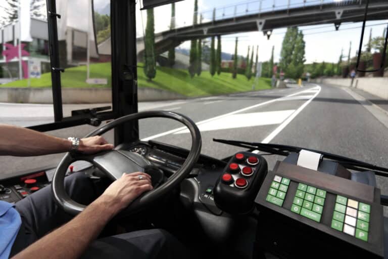Professional truck driver's perspective from the cab, hands on steering wheel navigating a highway with green landscape and overpass visible ahead.