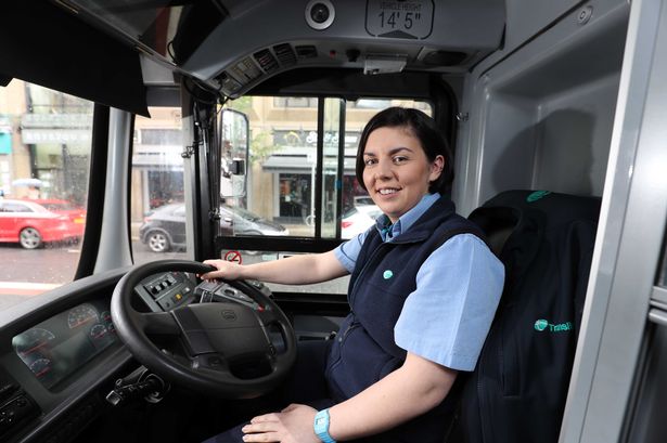 Female bus driver smiling at the wheel wearing a blue and navy uniform inside a transit bus during daytime.
