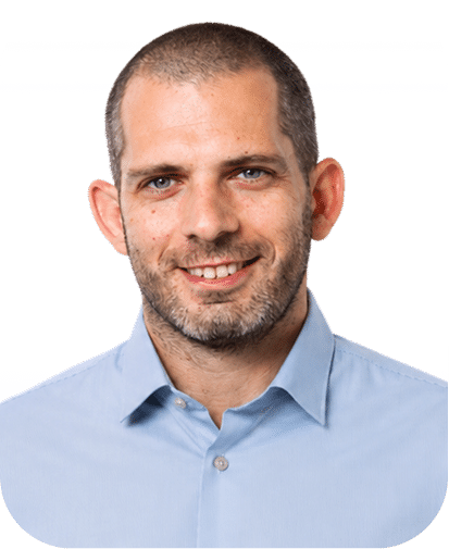 Professional headshot of a smiling man with short dark hair and facial stubble wearing a light blue button-up shirt against a white background.